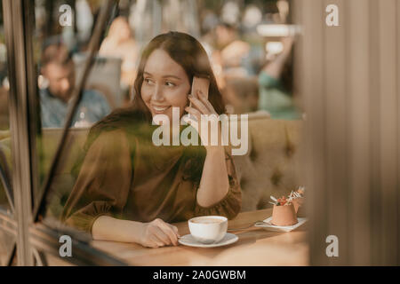 Charming brunette woman with long curly hair sitting at window in cafe with mobile phone in hands Stock Photo