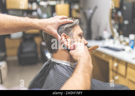 Making hair look magical. Side view of young bearded man getting shaved with straight edge razor by hairdresser at barbershop Stock Photo