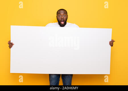 Handsome manly amazed mulatto guy showing, holding big wide copy space board, opened mouth, isolated over bright vivid yellow background Stock Photo