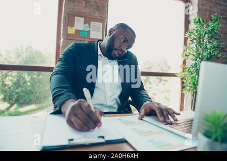 Handsome serious business shark, clerk, assistant mulato man in jacket, talking on phone, writing schedule at work place, workstation Stock Photo