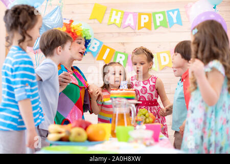 Preschool children holding by hands and blowing candle up. Little kid girl 5 years celebrating her birthday together with friends and clown Stock Photo