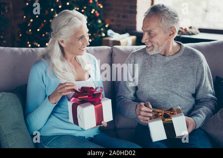 Portrait of nice sweet tender lovely charming cheerful wondered grey-haired married spouses wife husband sitting divan couch sofa in house opening bow Stock Photo