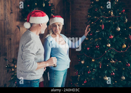 Two cheerful nice lovely sweet people grey-haired married spouses husband and wife grandma granddaddy helping each other decorating eve noel fir tree Stock Photo