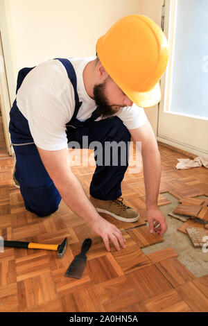 Construction worker and constructor is working on renovation of apartment. Handyman removing parquet flooring using yellow hammer and scraping tool. Stock Photo