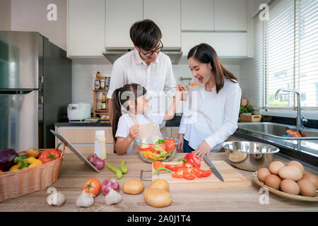 Asian daughters feeding salad to her mother and her father stand by when a family cooking in the kitchen at home.  Family life love relationship, or h Stock Photo