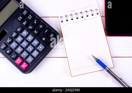 Blank note book with pen beside are smart phone,calculator, glasses and on white wooden table, business concept. Stock Photo