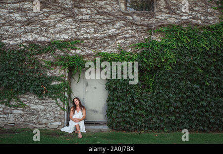 Woman in a white dress sitting in the doorway of an ivy covered wall. Stock Photo