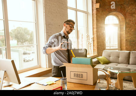 Young businessman fired, looks angry. Has to pack his office belongings and to leave work place for new worker. Problems in occupation, stress, unemployment, new way of life or end of career. Stock Photo