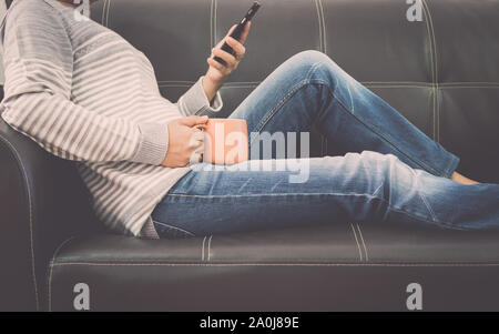 Woman is on leather sofa using mobile phone and holding a coffee cup at home. Stock Photo
