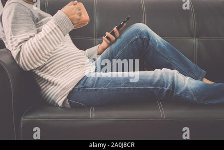 Woman is on leather sofa using mobile phone and holding a coffee cup at home. Stock Photo