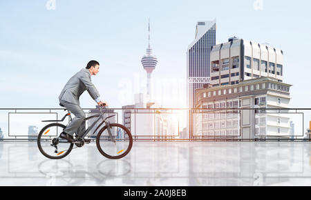 Man wearing business suit riding bicycle on penthouse balcony. Handsome cyclist on background of city center architecture. Terrace with modern downtow Stock Photo