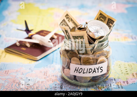 Holidays budget concept. Holidays money savings in a glass jar with compass, passport and aircraft toy on world map Stock Photo
