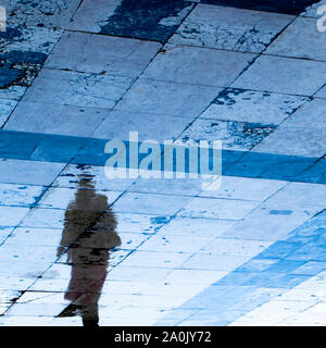 Blurry reflection shadow silhouette on wet city sidewalk of a mysterious person walking away alone in the night Stock Photo