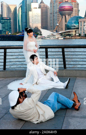 A photographer lies on the ground to photograph a wedding couple in formal attire posing at The Bund with Financial District in background, Shanghai,