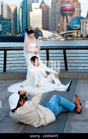 A photographer lies on the ground to photograph a wedding couple in formal attire posing at The Bund with Financial District in background, Shanghai, Stock Photo