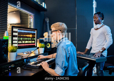 Young man recording music by computer while African guy playing pianoboard Stock Photo