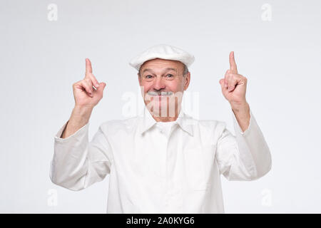 Smiling senior cook in white uniform smiling and pointing up trying to pay attention to important information. Stock Photo
