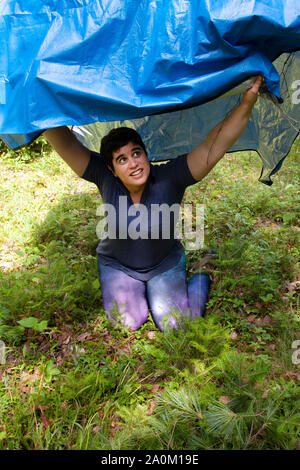 person takes shelter under a blue tarp in the forest while camping Stock Photo