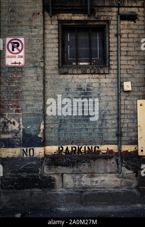 A weathered brick wall in an alleyway featuring a no parking sign a barred basement window and peeling paint with a gritty urban aesthetic Stock Photo