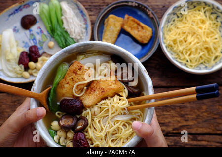 Close up woman hand hold Vietnamese vegan noodles soup on ingredients food background, vegetarian dish with bok choy, egg noodle, lotus seed, tofu Stock Photo