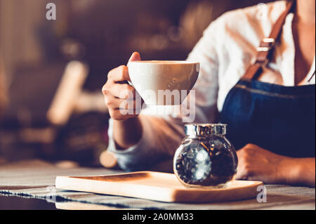 Closeup of professional female barista hand making and holding white cup of coffee. Happy young woman at counter bar in restaurant background. People Stock Photo
