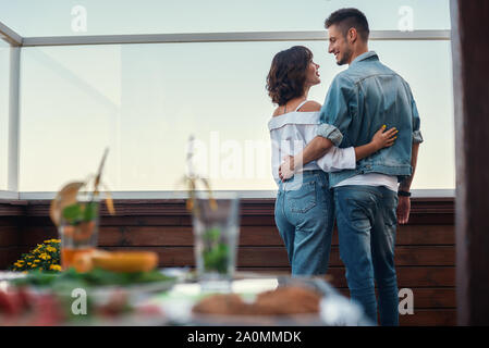 Romantic evening. Back view of young couple in casual clothes are looking at each other while standing on rooftop terrace. Relationship concept Stock Photo