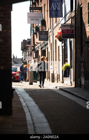 A woman (age 30s) walks towards purposefully towards Leen Lane, a small lane overshadowed by buildings in central Chester. An open sunlit area behind. Stock Photo