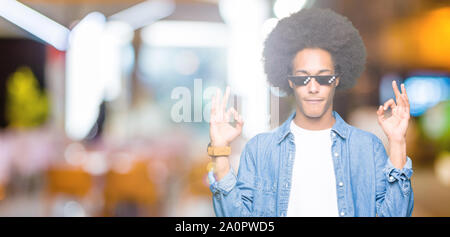 Young african american man with afro hair wearing thug life glasses relax and smiling with eyes closed doing meditation gesture with fingers. Yoga con Stock Photo