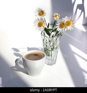 A cup of black coffee with foam, a bouquet of white camomile flowers in a crystal vase with water on a white table in the sunlight and shadow Stock Photo
