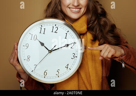 Closeup on smiling trendy woman in yellow beret and scarf pointing at round clock remind about autumn time change on brown background. Stock Photo