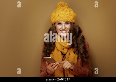 smiling trendy woman in yellow beret and scarf with smartphone hanging in social media and against bronze background. Stock Photo