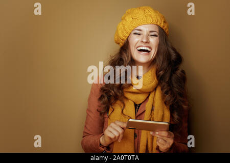 Portrait of smiling young woman in yellow beret and scarf with phone and texting on brown background. Stock Photo