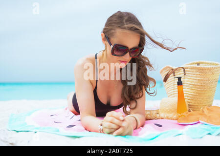 pensive trendy middle age woman with long curly hair in elegant black bathing suit on a white beach laying on a round towel and sun tanning. Stock Photo