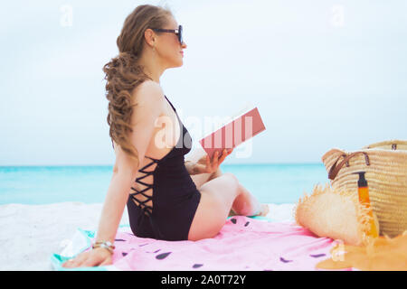 trendy 40 year old woman with long curly hair in elegant black swimsuit with book looking into the distance on a white beach. Stock Photo