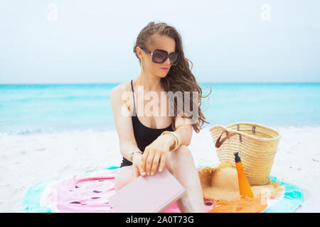 pensive stylish middle age woman with long curly hair in elegant black bathing suit with book sitting on round watermelon towel on a white beach. Stock Photo