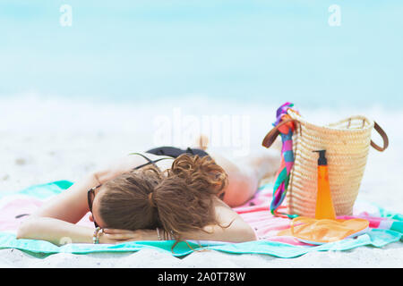 trendy 40 year old woman with long curly hair in elegant black bathing suit on a white beach sleeping while sun bathing. Stock Photo
