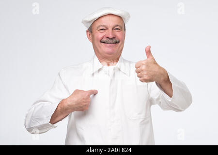 mature hispanic chef cook showing thumb up and pointing at his chest being proud on white background. Studio shot Stock Photo