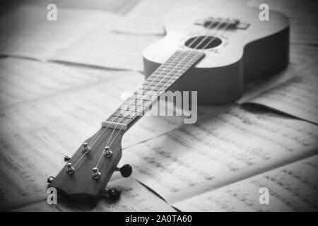 A black-and-white image of a ukulele lying on scattered sheets with musical notes left by the composer. Stock Photo