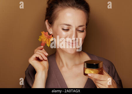 Hello autumn. Portrait of smiling modern woman in a bathrobe with autumn leaf earring looking at facial creme isolated on bronze background. Stock Photo