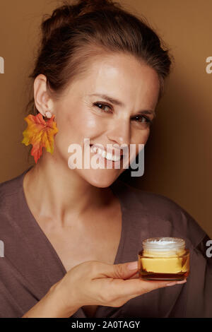 Hello autumn. Portrait of happy modern 40 year old woman in a bathrobe with autumn leaf earring holding facial creme isolated on bronze background. Stock Photo