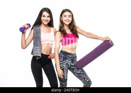 Photo of two attractive multiethnic sportswomen in tracksuits smiling and holding yoga mat while standing over white wall outdoors Stock Photo