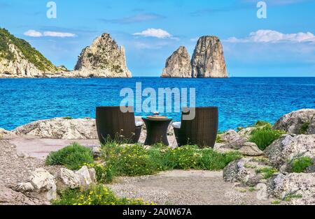 An outdoor table and two chairs on the water's edge, with a dramatic view of the famous limestone pinnacle rocks of Capri, known as the Faraglioni. Stock Photo