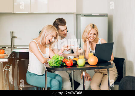 Three students in domestic clothes sitting at the table in kitchen, going to prepare breakfast, watching video recipe on laptop, drinking orange juice Stock Photo