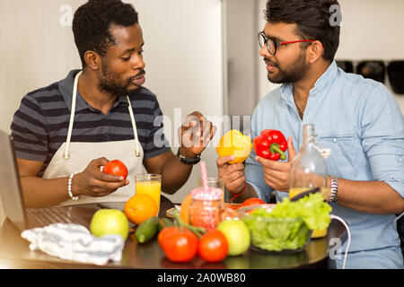 Closeup shot of two multi ethnic bearded friends in casual clothes preparing vegetable salad or smoothie, sitting at table talking using laptop comput Stock Photo
