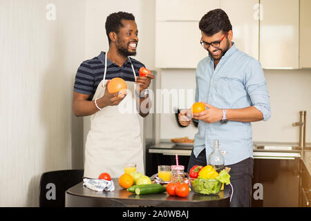 Two male students in casual clothes going to cooking smoothie standing in kitchen of hostel, holding grapefruit, orange, apple, tomato in hands, cheer Stock Photo