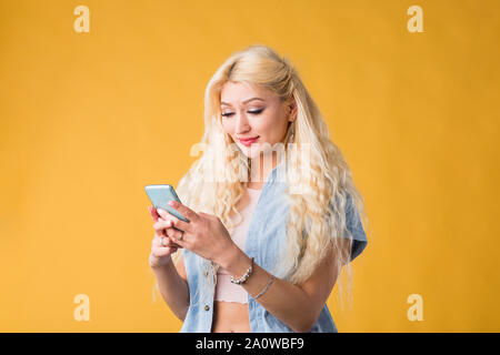 Lovely caucasian serious student with long hair, looking at screen of cell phone, making video call, reading news, using wireless internet on yellow b Stock Photo