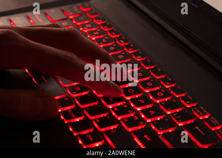 Close up of fingers typing on glowing red backlight keys on a computer keyboard. in the dark. Computer hacker, internet fraud or spying concept. Stock Photo