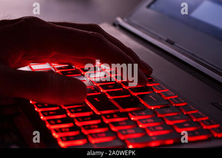 Close up of fingers typing on glowing red backlight keys on a computer keyboard. in the dark. Computer hacker, internet fraud or spying concept. Stock Photo