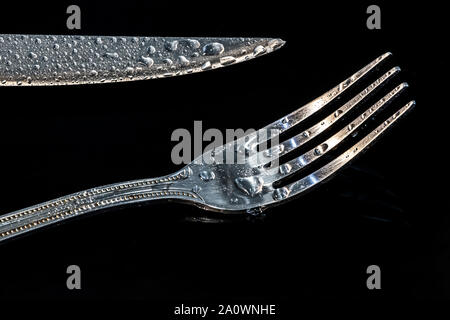 Fork and knife at the black table with water droplets on it. Conceptual image about danger of eating too much tasty food. Stock Photo