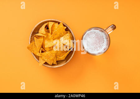 top view of crispy nachos in wooden bowl near glass of beer on orange background, Mexican cuisine Stock Photo